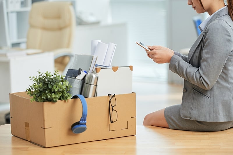 woman-suit-sitting-desk-office-with-belongings-box-using-smartphone
