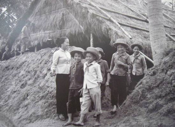 Cô giáo tươi cười đứng trước cửa lớp chỉ dẫn học sinh khi tan học. Các học trò đầu đội mũ rơm, miệng tươi cười ra về.