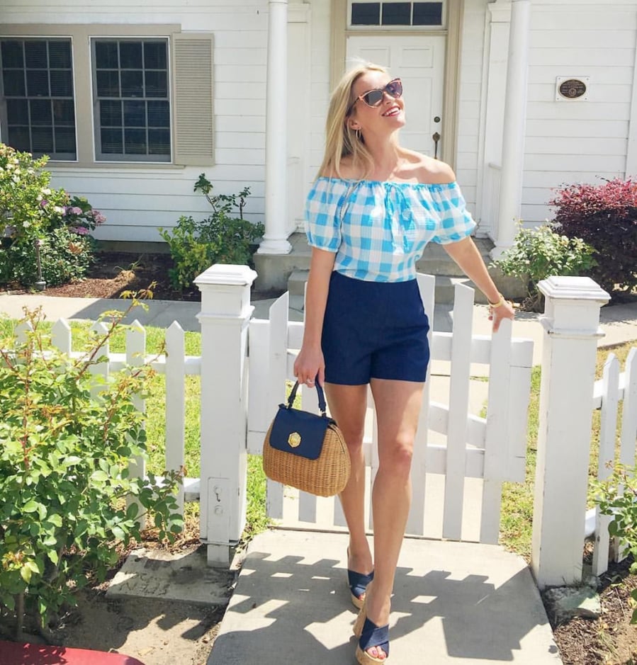 Reese Witherspoon chọn phong cách đơn giản, thoải mái với áo mi trễ vai kẻ ô và quần short cạp cao kết hợp cùng túi xách cói đan kiểu cách.